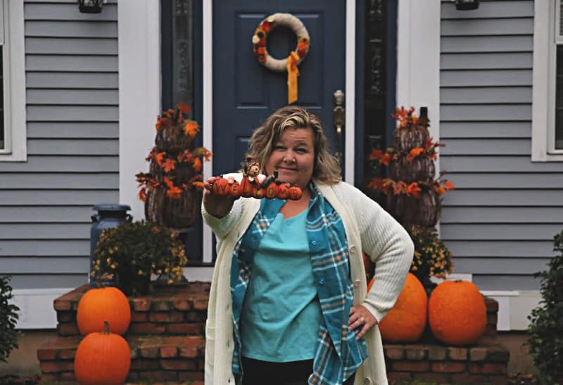 woman in plaid flannel tunic, tank, cream color quilted car coat holding fall theme welcome sign 