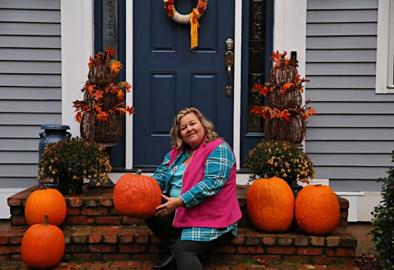 woman in pink fleece vest, flannel tunic and dark denim holding pumpkin sitting on stairs 