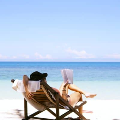 woman with beach hat sitting in chair reading on beach