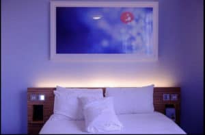 Bedroom with white bedding on bed , backlit headboard and soothing art on wall above bed 