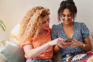 two women sitting close together on a couch looking at one of their phone screens together