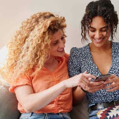 two women sitting close together on a couch looking at one of their phone screens together