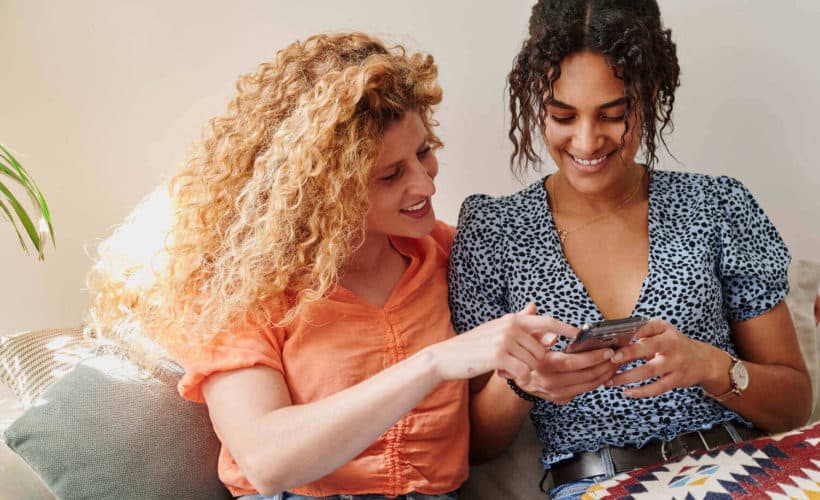 two women sitting close together on a couch looking at one of their phone screens together