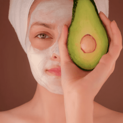 closeup of woman holding half an avocado in front of half of her face, with the other half covered in a beauty mask