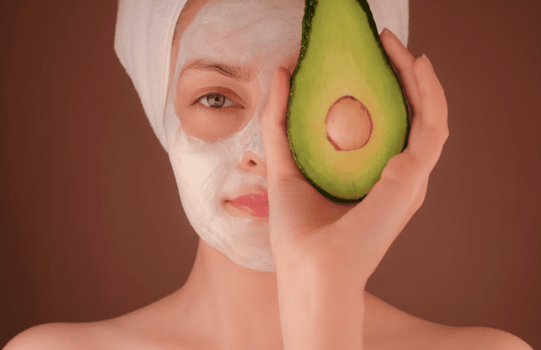 closeup of woman holding half an avocado in front of half of her face, with the other half covered in a beauty mask