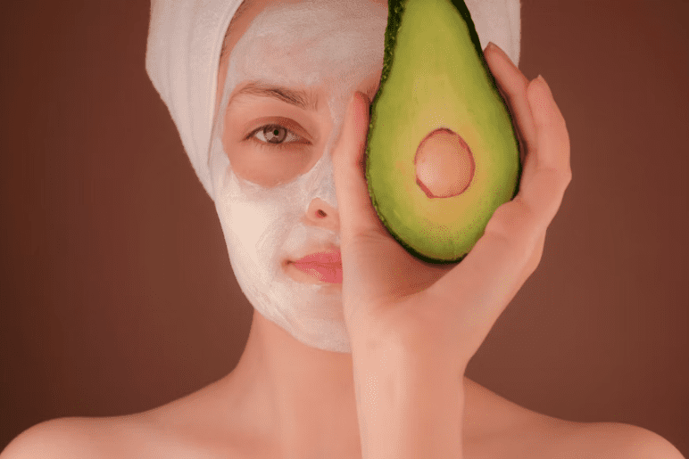 closeup of woman holding half an avocado in front of half of her face, with the other half covered in a beauty mask